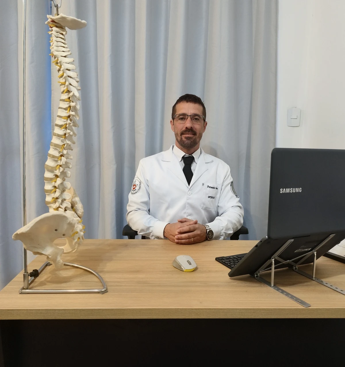 Dr. Fernando Guerra sentado à mesa do consultório, com um modelo de coluna vertebral ao lado