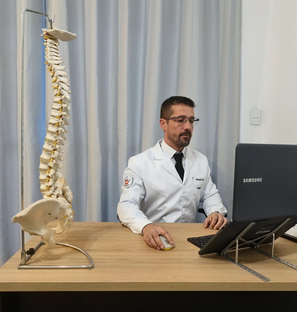 Dr. Fernando Guerra sentado à mesa do consultório, ao lado de um modelo de coluna vertebral