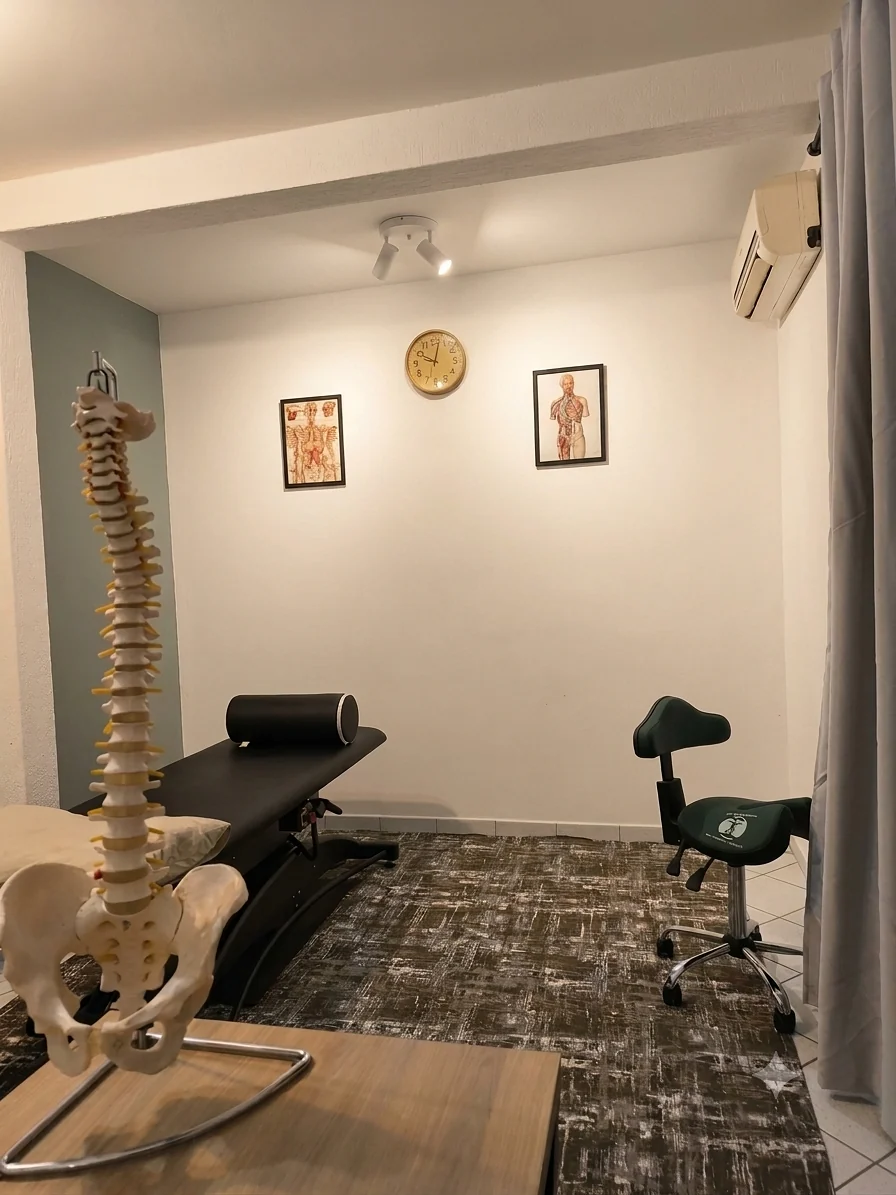 Sala de atendimento com maca preta, modelo de coluna vertebral e quadros de anatomia